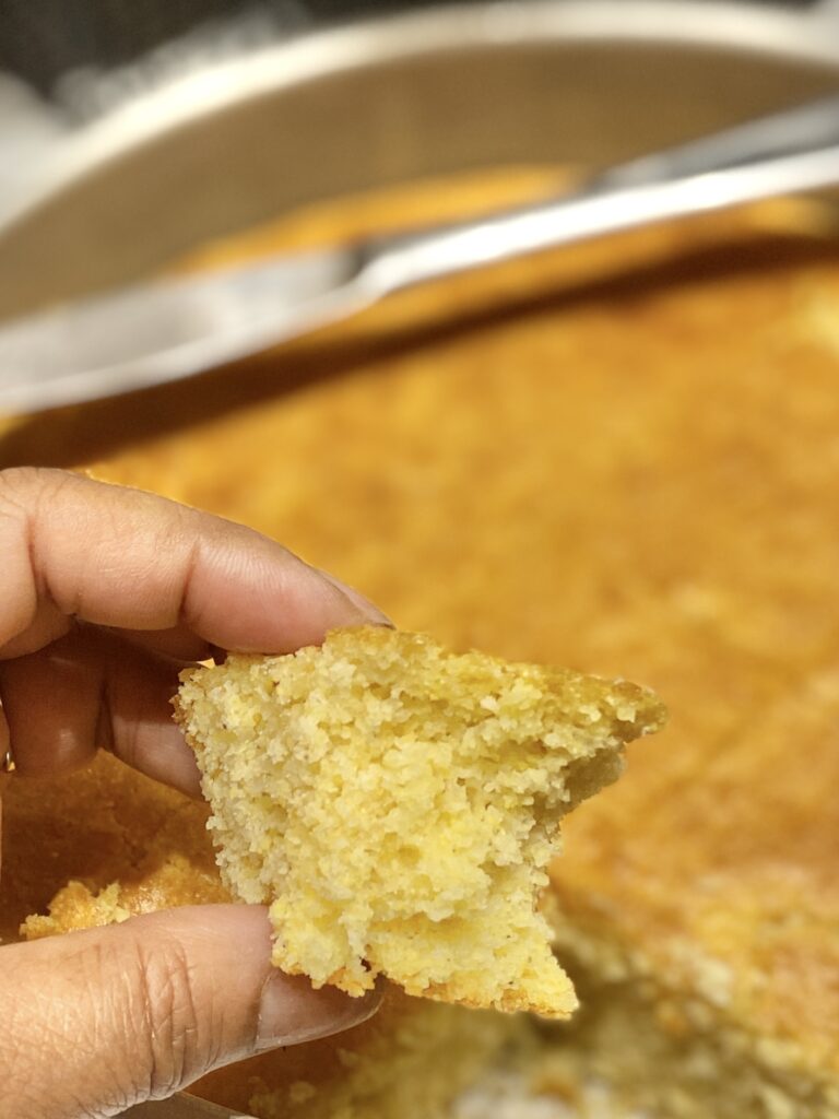 a woman holding a small piece of egg free cornbread in front of a large pan of cornbread