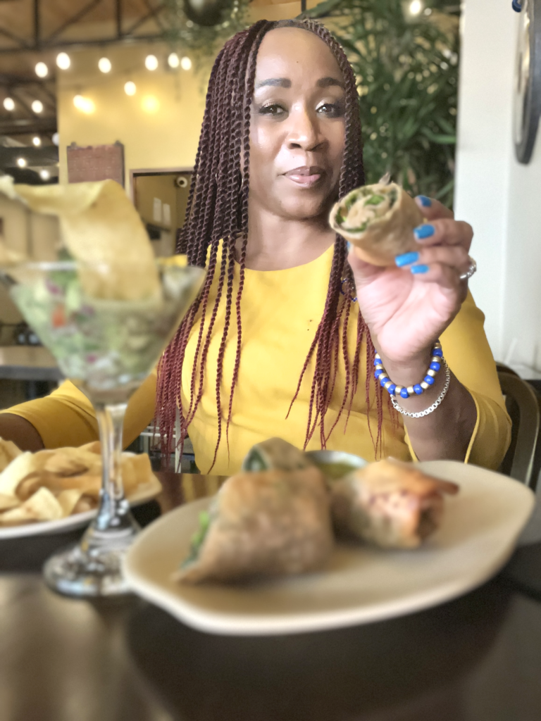 woman in yellow holding up a tex mex egg roll with coconut ceviche in a martini glass in front of her